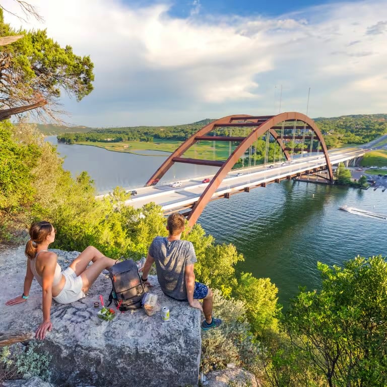 Two people sitting on a rock overlooking an urban bridge over a river