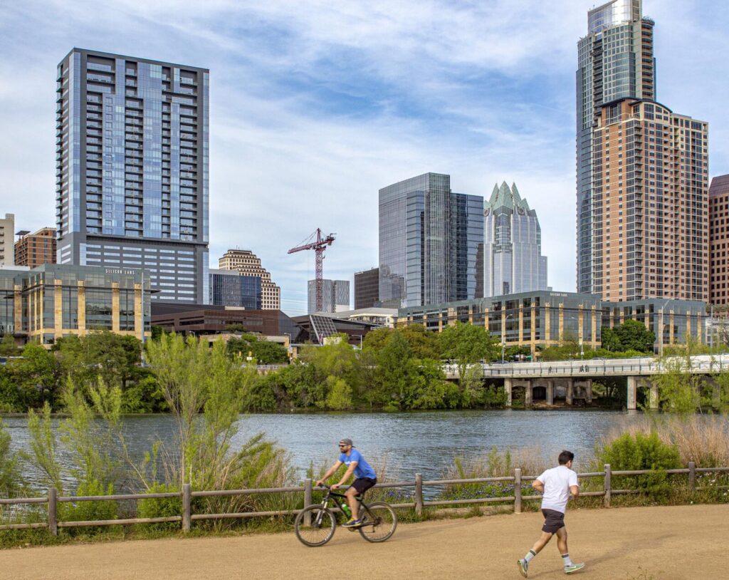 A person biking and a person running on a path in front of a river and skyscrapers