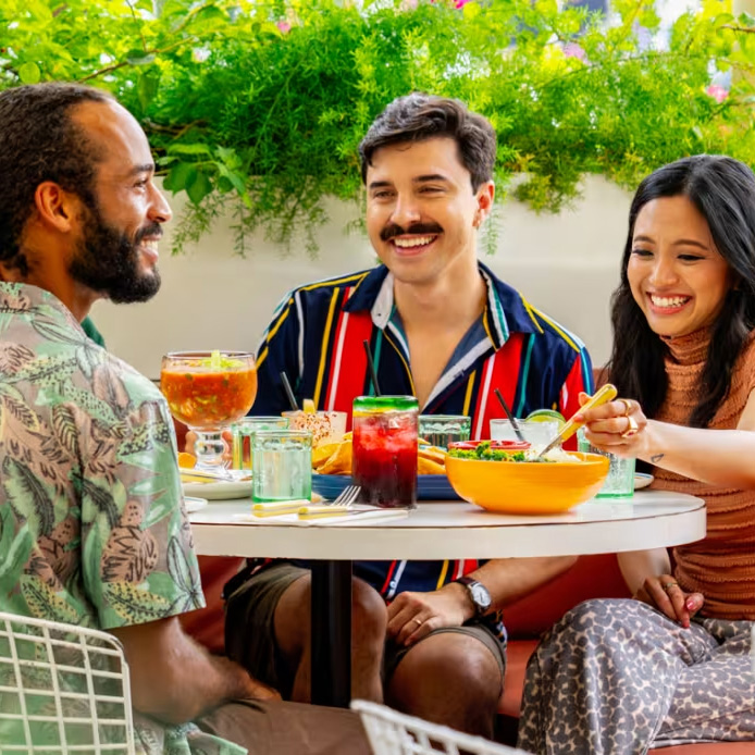 Three young people sitting at an outdoor table filled with food and drink