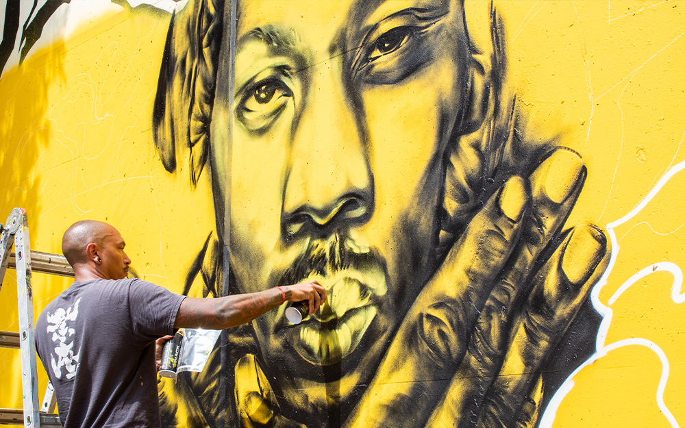 A man spray painting an image of Tupac on a yellow concrete wall
