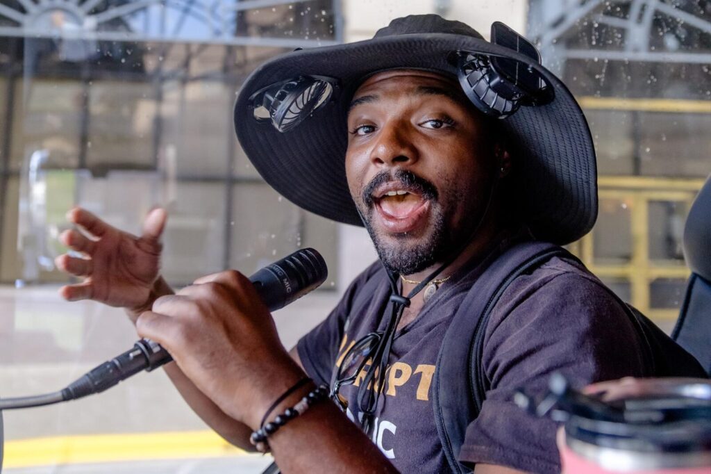 A man with a fan-hat speaking into a microphone on a bus